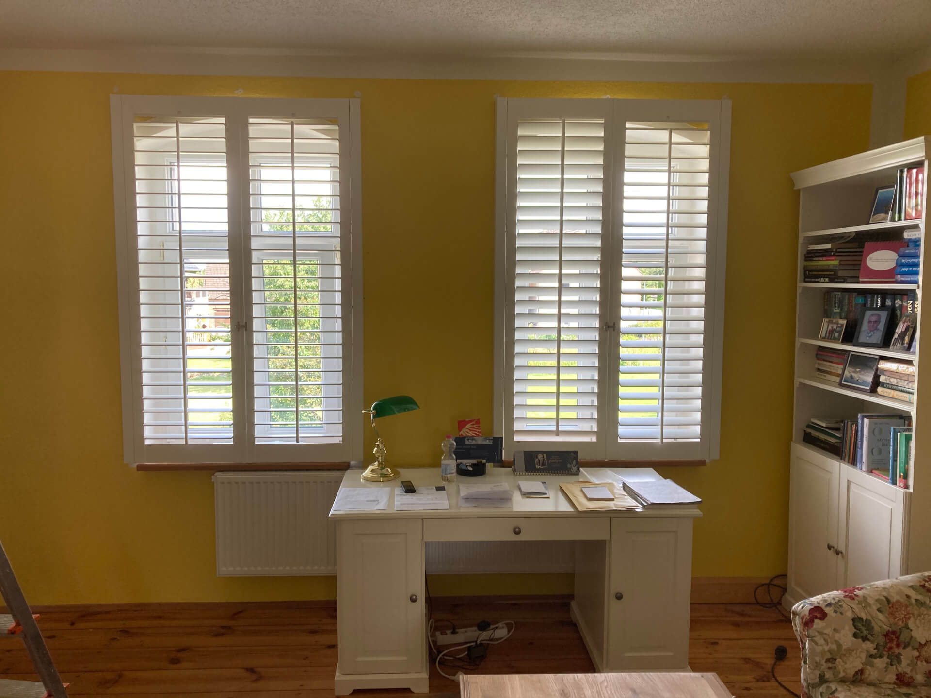 Arbeitszimmer mit gelben Wänden und zwei großen Fenstern, die mit weißen Lamellen-Fensterläden ausgestattet sind. Vor den Fenstern steht ein klassischer weißer Schreibtisch mit einer grünen Schreibtischlampe und Schreibutensilien. Rechts befindet sich ein