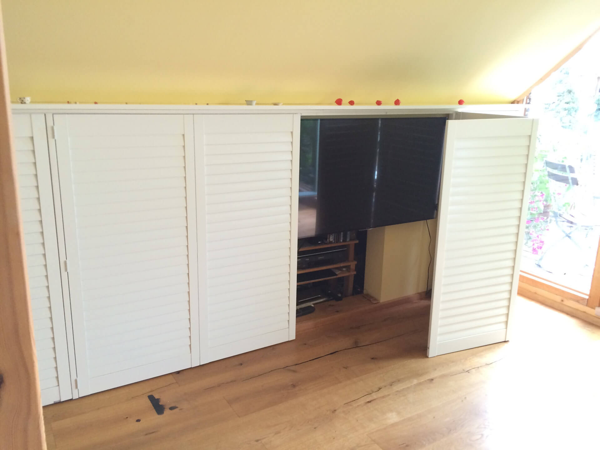 Dachgeschoss mit eingebauter Stauraumlösung aus weißen Lamellentüren, hinter denen ein Fernseher und Elektronikgeräte versteckt sind, kombiniert mit einer warmen Holzbodenoptik und gelber Wand.