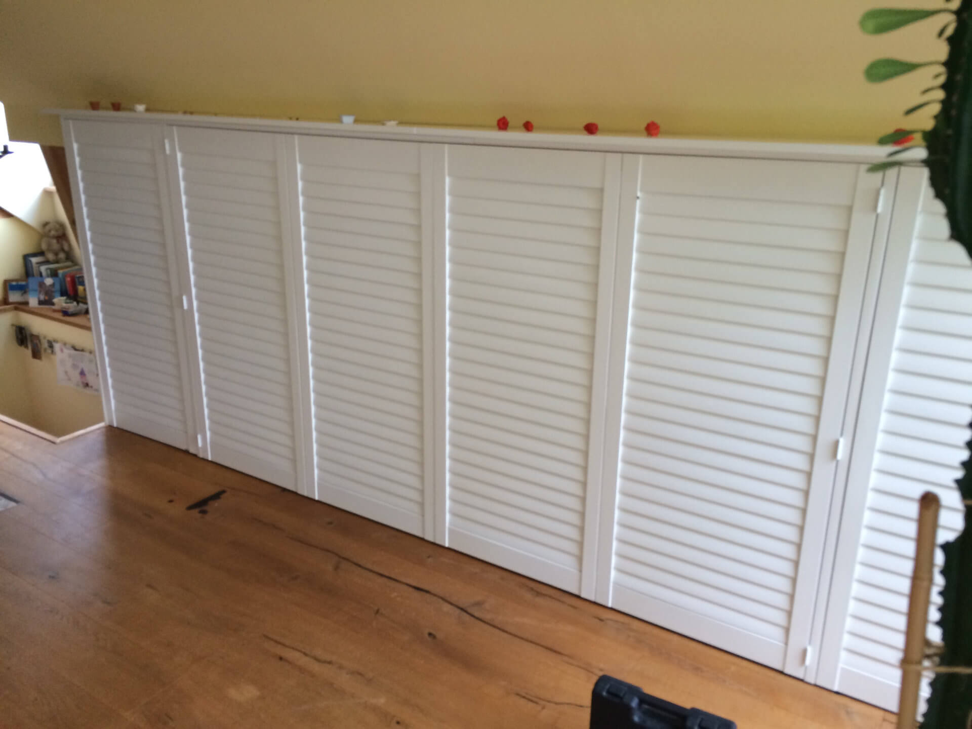 Dachgeschoss mit maßgefertigten weißen Lamellentüren als Stauraumlösung, kombiniert mit einer warmen Holzbodenoptik und einer schrägen gelben Wand.